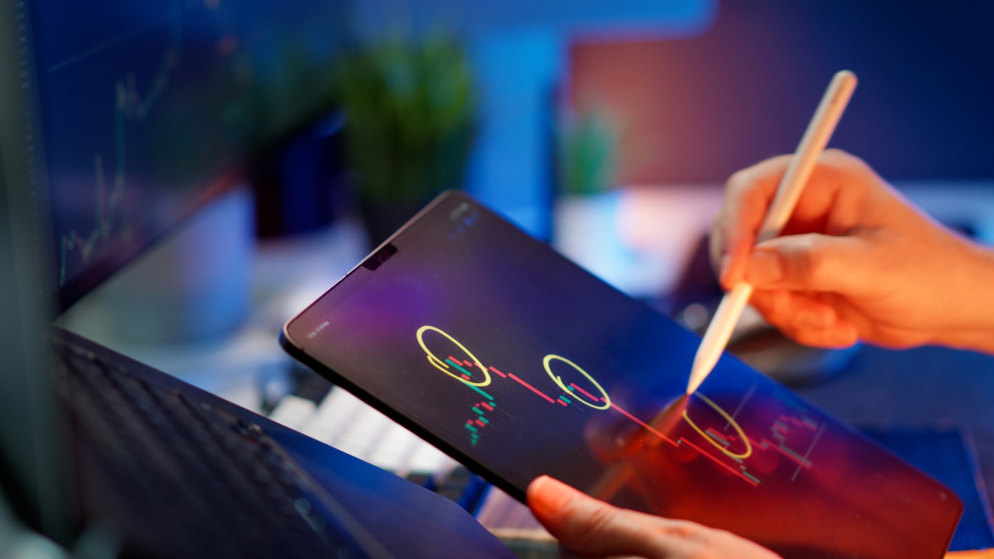 Hands use a digital stylus to analyze financial charts on a tablet.